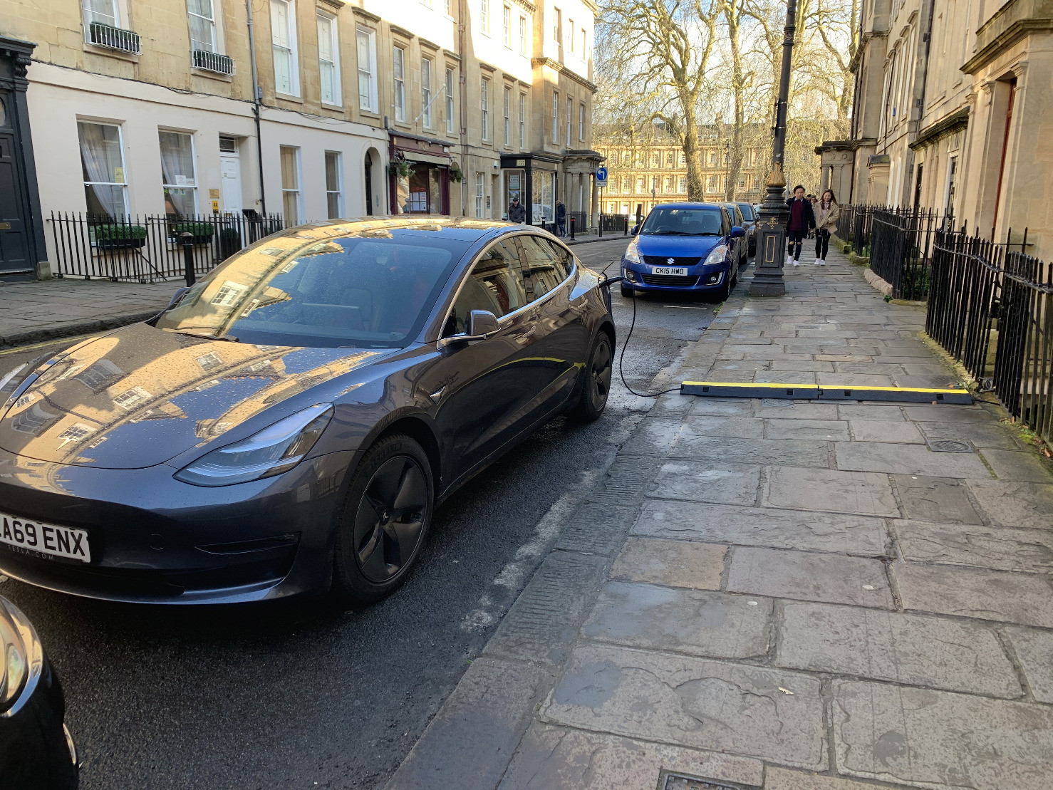 A Tesla parked outside of a house in bath. There’s a plastic cable protector over the pavement and the car is being charged. The protector is yellow on top but is pretty tall and easy to trip over.