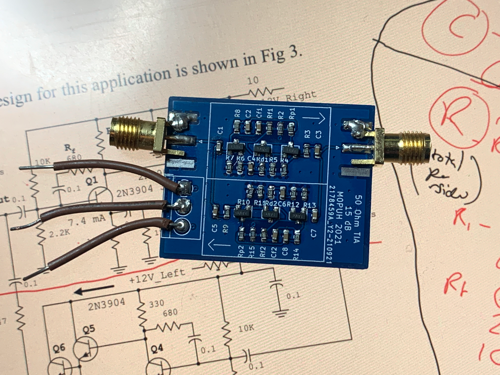 A small dark blue printed circuit board resting on an LCD display. The board has SMA connectors on the top left and right and three wires on the left. On the board are two almost identical circuits made of 0603 surface mount components and SOT 23 transistors. 