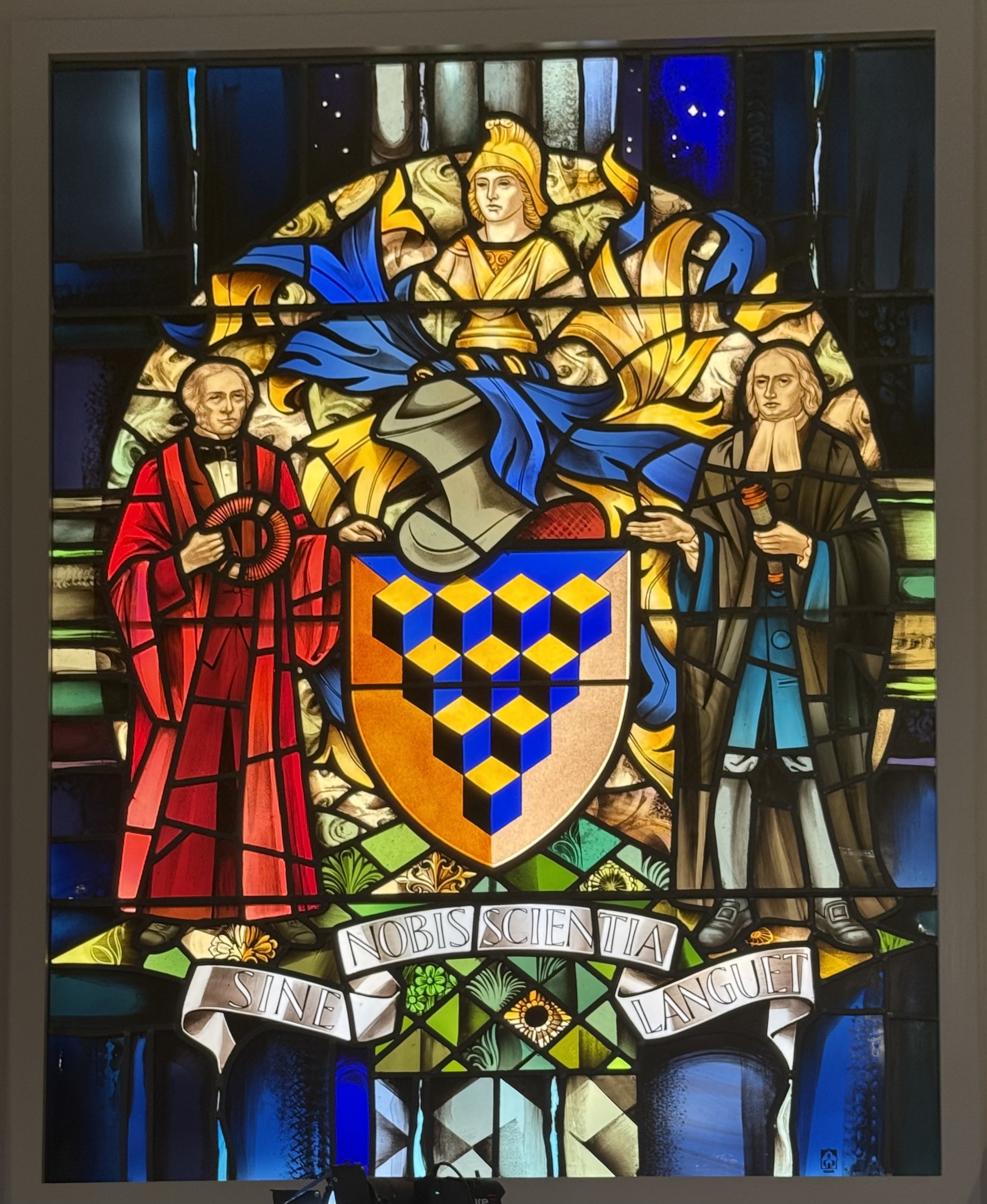 A stained glass coat of arms depicting a man in red robes on the left and a man in dark robes on the right. The man on the left is Faraday and is holding an inductor. The man on the right is Newton holding a telescope. In the centre at the top is a depiction of Minerva, in gold and in the centre under miners a is a knight’s helmet on top of a shield with a picture of a stack of blue and yellow cubes. Finally across the bottom is the motto “Sine Nobis Scientia Languet”