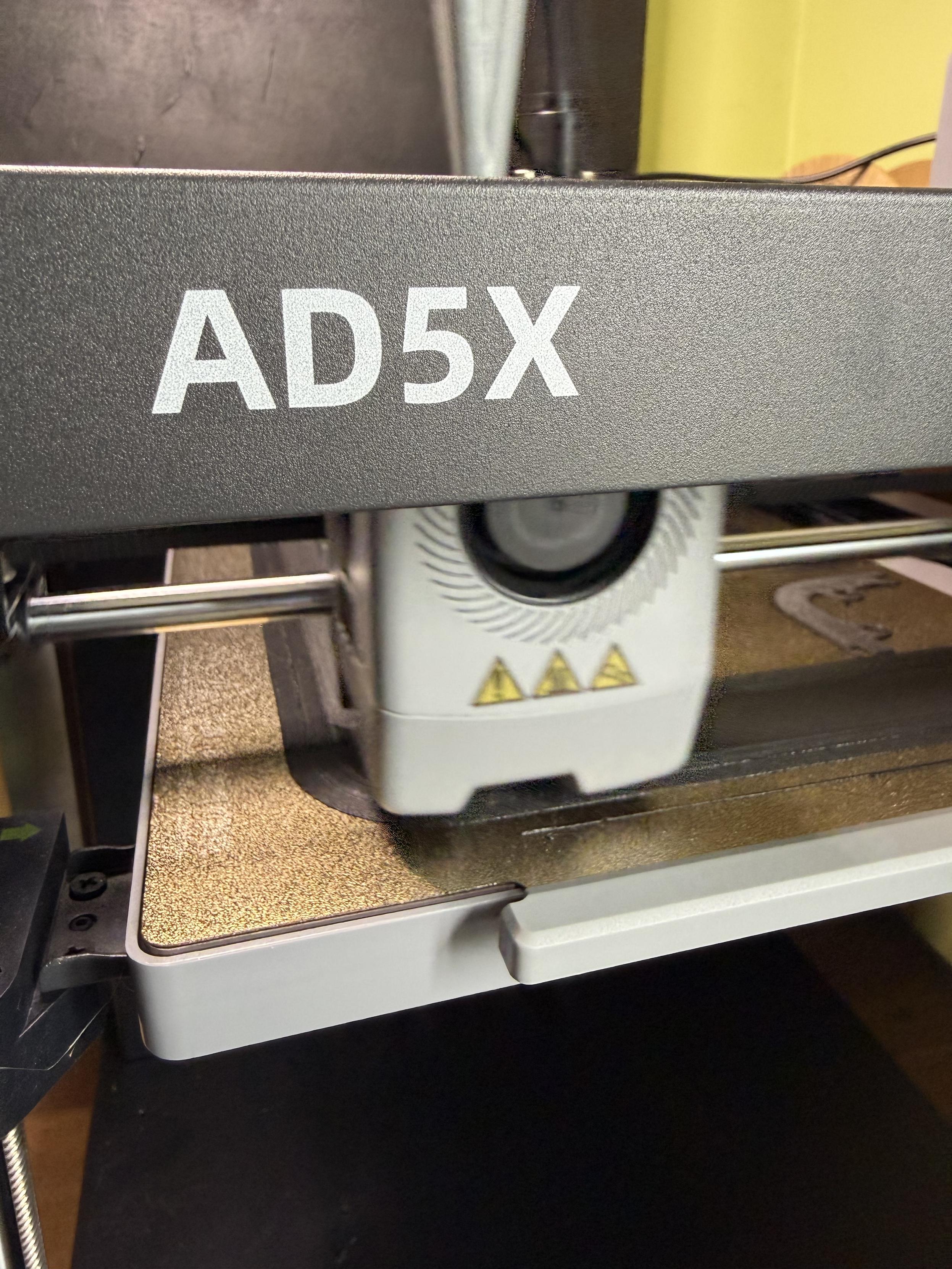 Close up of a 3D printer showing the AD5X label and the head, which is just behind and slightly motion blurred.